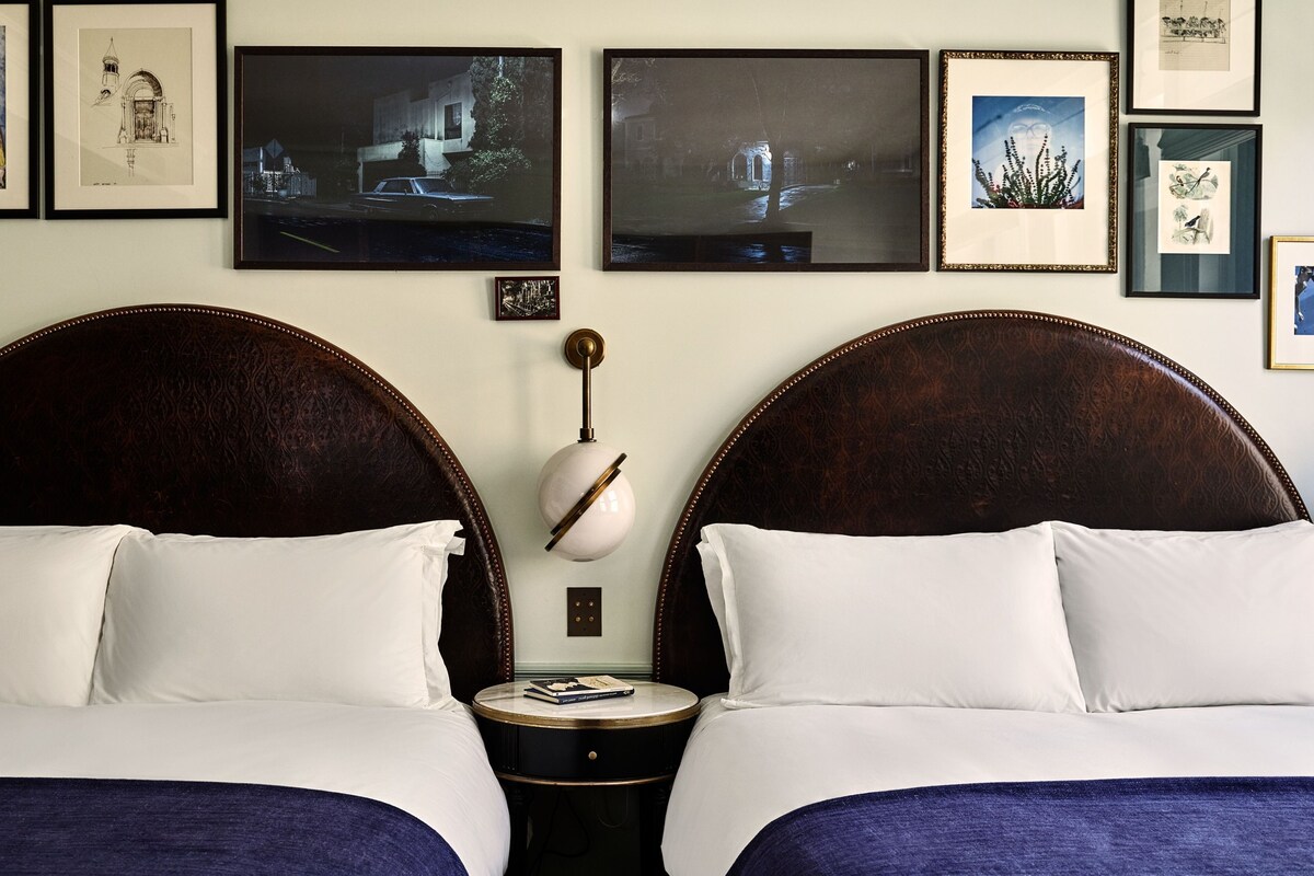 A hotel room with two beds, a round table with a lamp, and framed artwork on the wall.