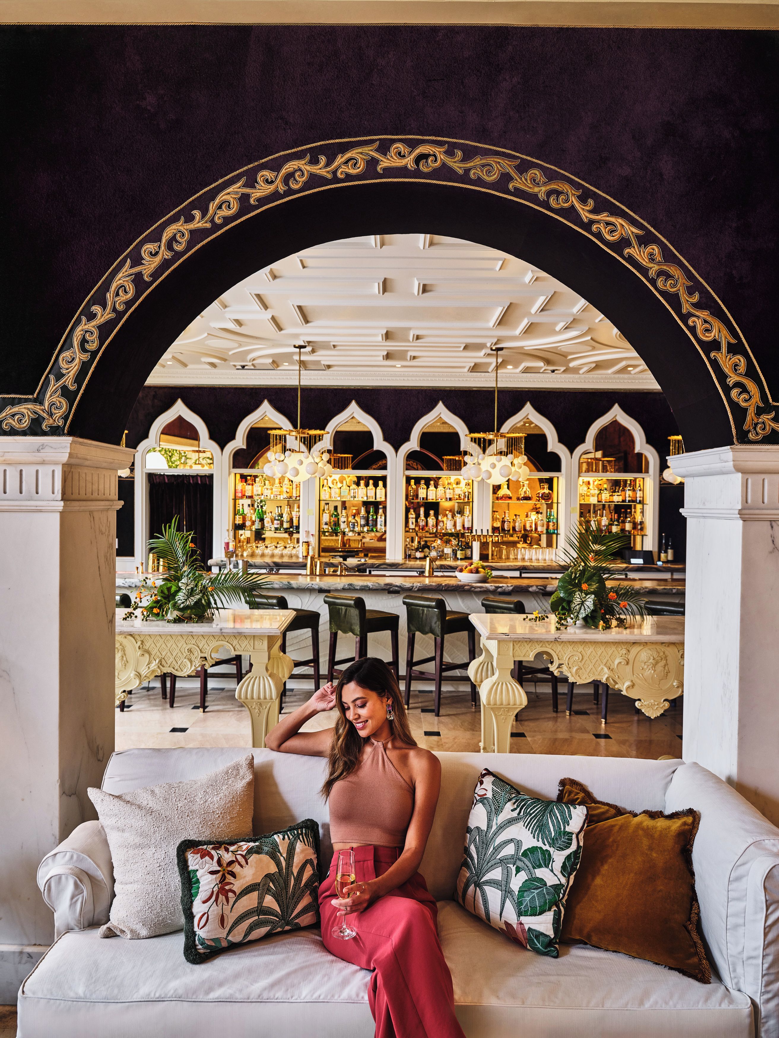 A woman seated on a sofa with a drink, in a room with a bar and ornate ceiling.