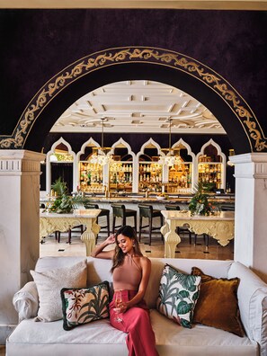 A woman seated on a sofa with a drink, in a room with a bar and ornate ceiling. - Hotel Per La, Autograph Collection (Los Angeles)
