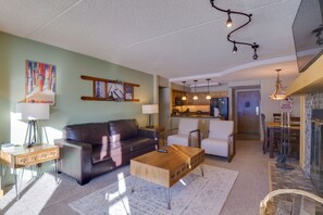 A living room with a brown leather couch, a wooden coffee table, and a white armchair. There is a kitchen area visible in the background.