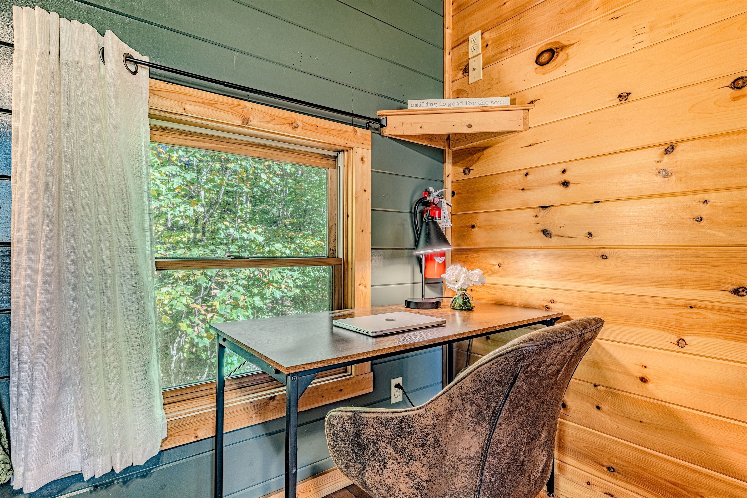 A wooden cabin interior with a desk, chair, window with curtains, and a shelf with a sign.