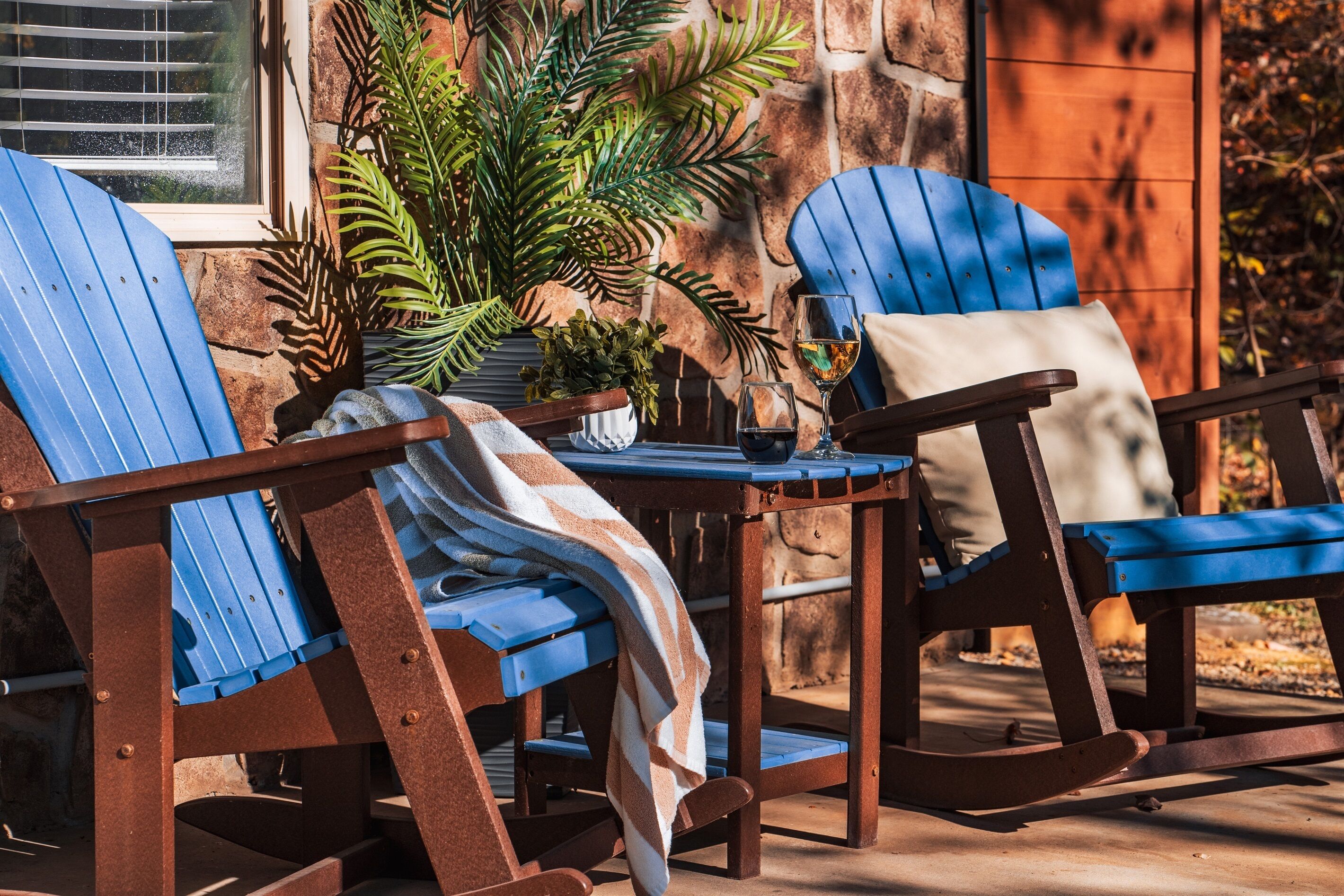 A patio with two rocking chairs and a table, surrounded by potted plants and a stone wall.