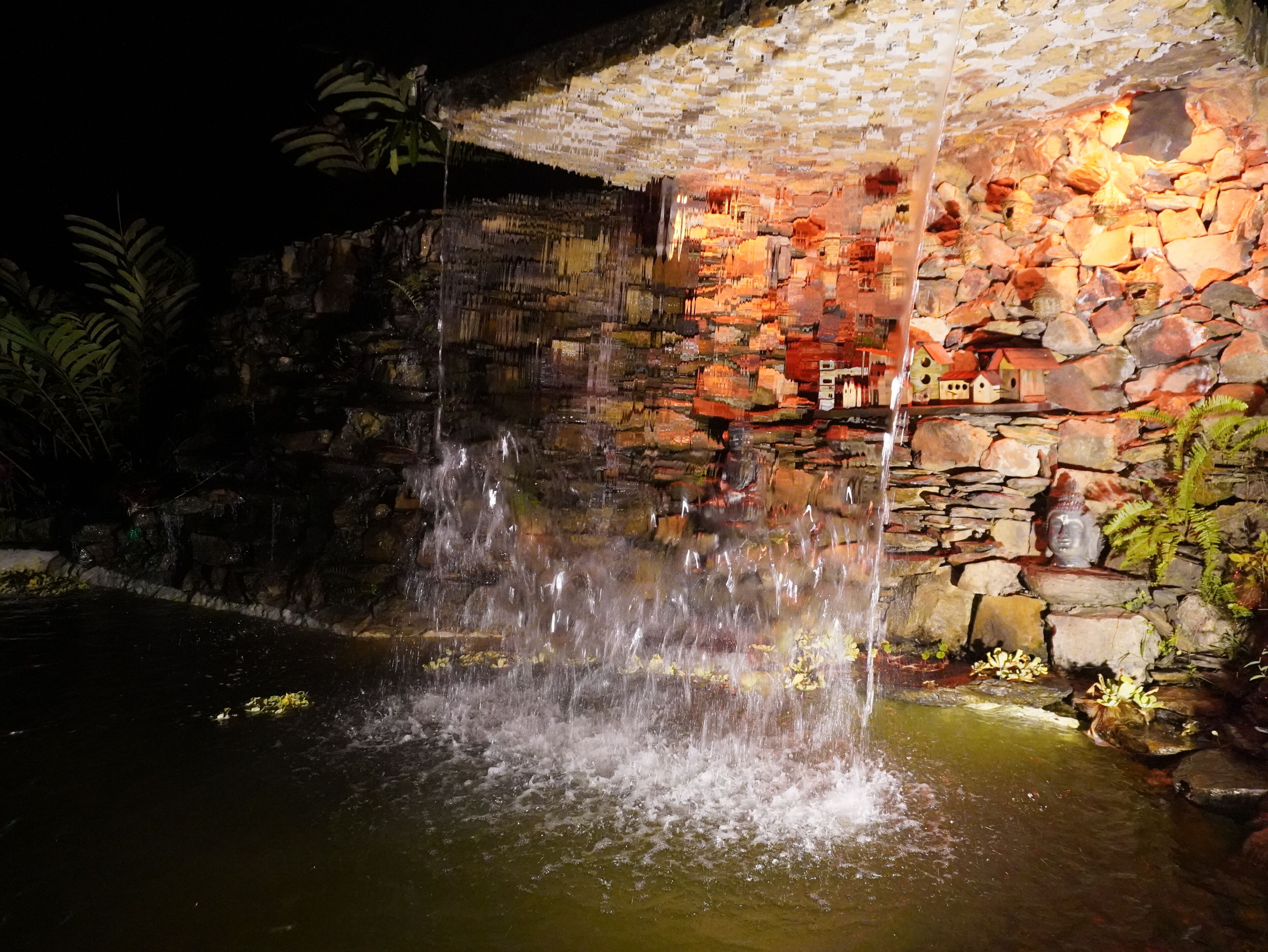 A nighttime waterfall feature with a stone wall backdrop and a Buddha statue.