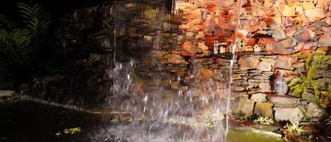 A nighttime waterfall feature with a stone wall backdrop and a Buddha statue.