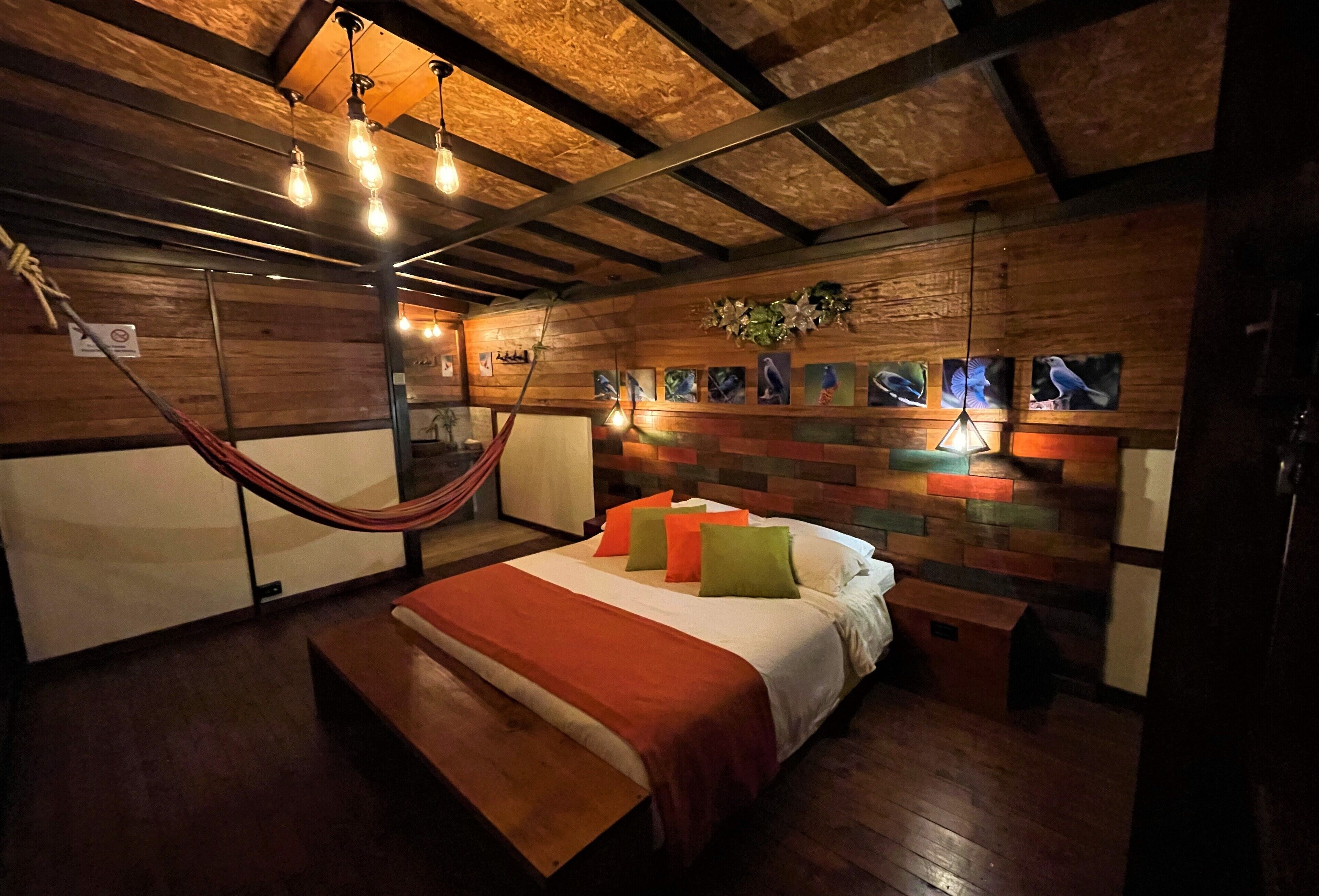 Un dormitorio con techo de madera, cama con almohadas anaranjadas y verdes, y una hamaca.