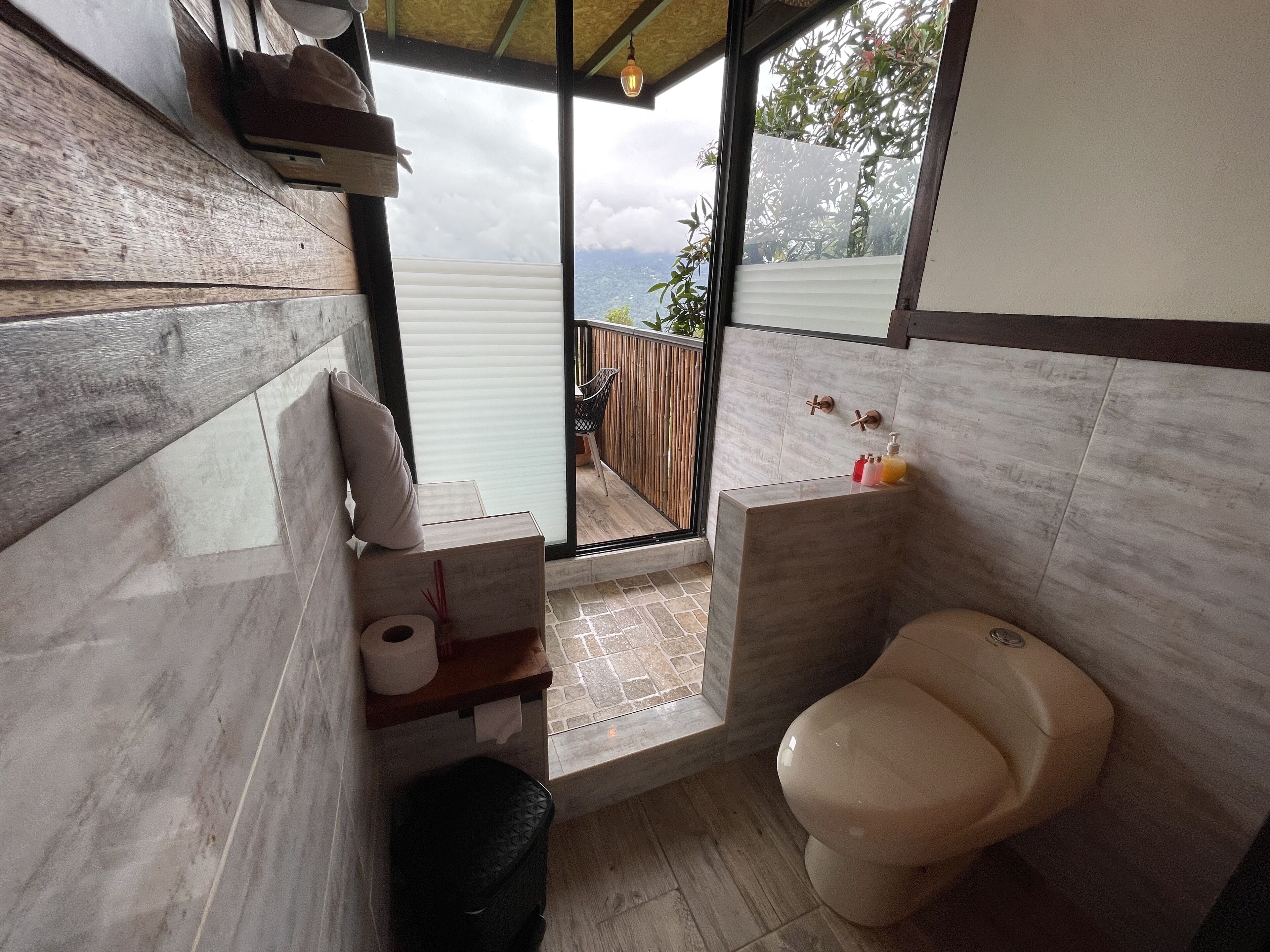 A modern bathroom with a toilet, a sink, and a small shelf with toiletries. There is a window providing a view of the outdoors.