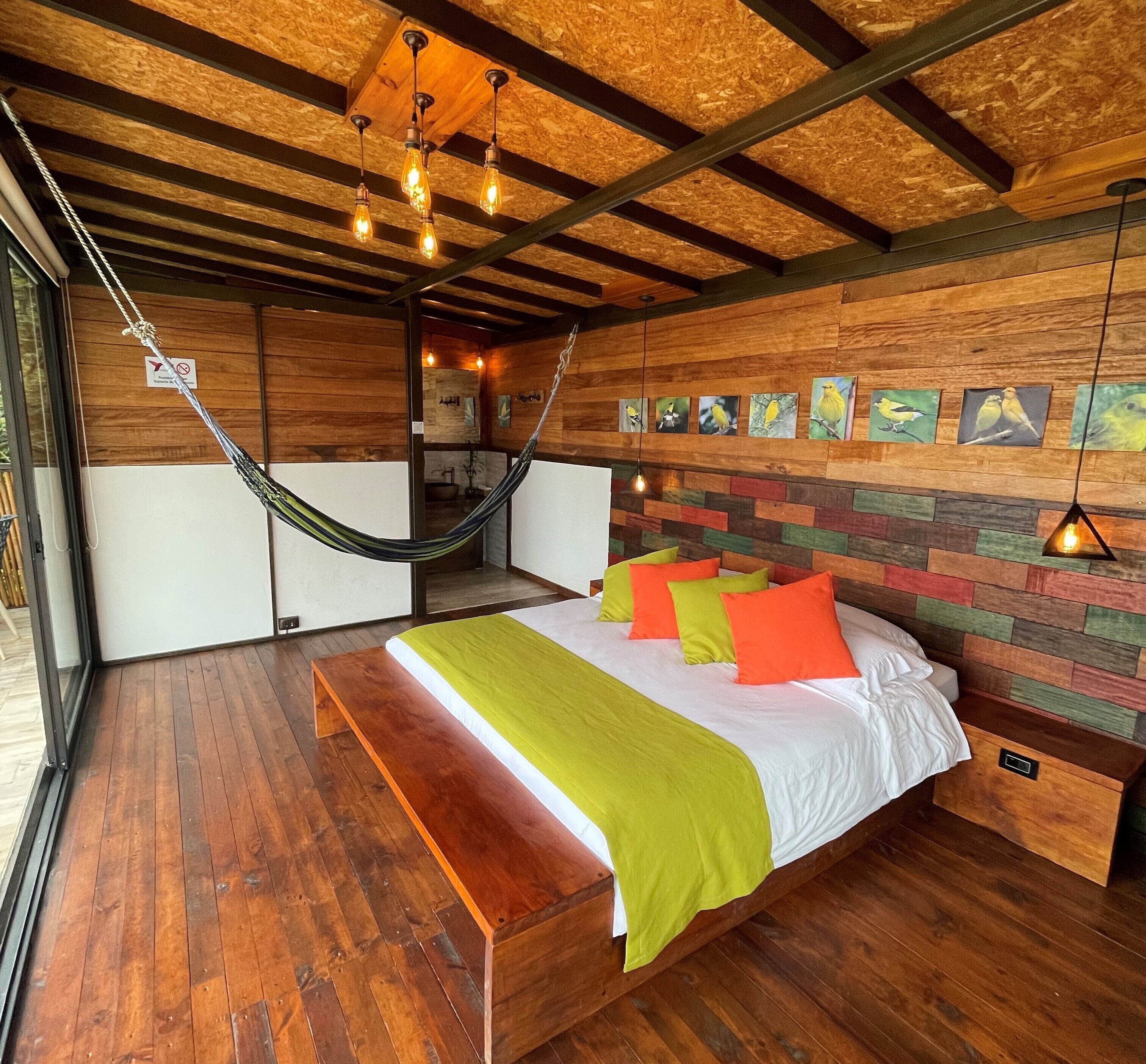 Un dormitorio con piso de madera, cama con cubrecama verde y blanco, almohadas naranja y verde, una hamaca y una pared con pinturas de pájaros.