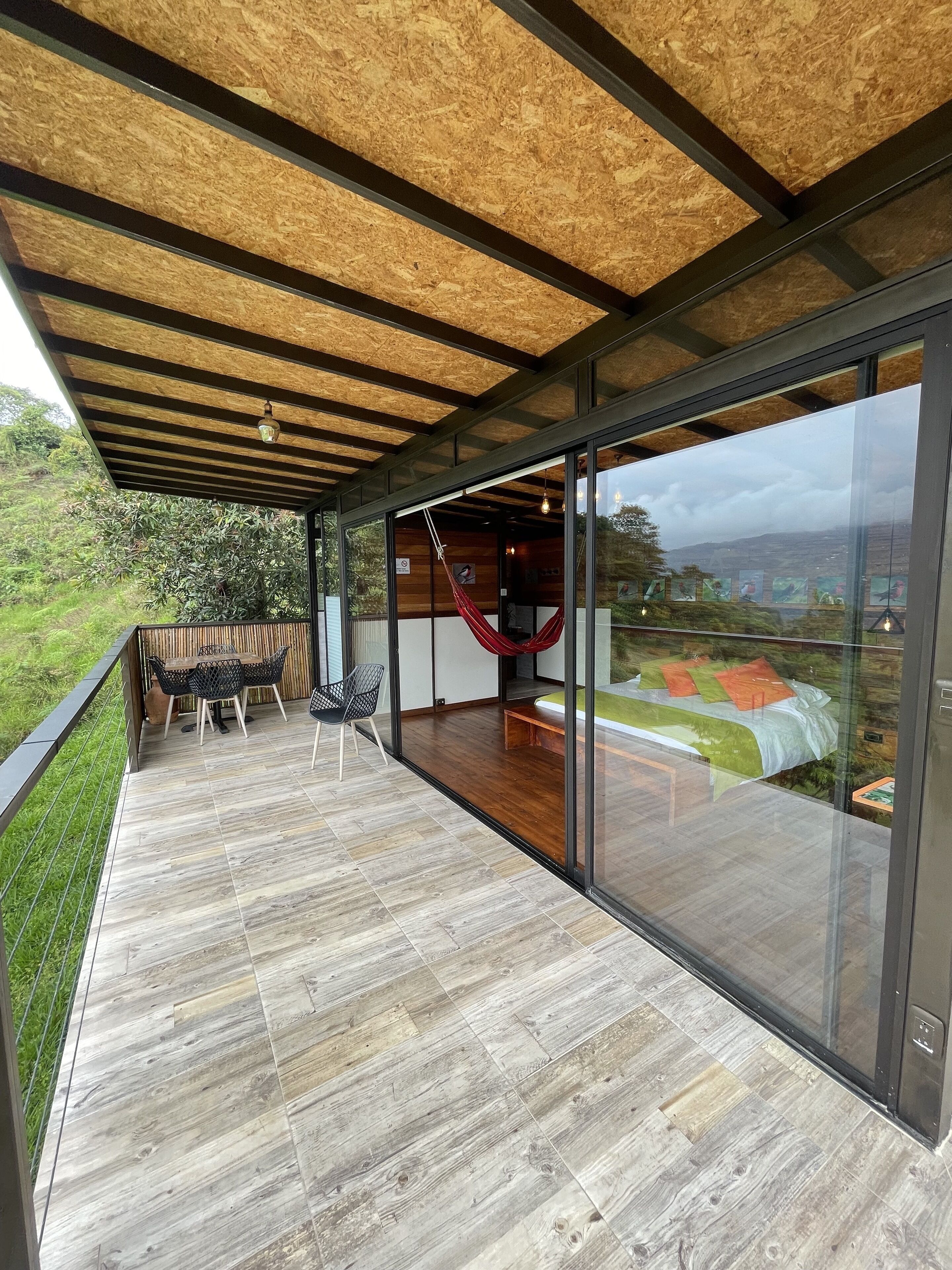 Een modern terras buiten met een houten vloer, een hangmat en een glazen wand met uitzicht op het landschap.
