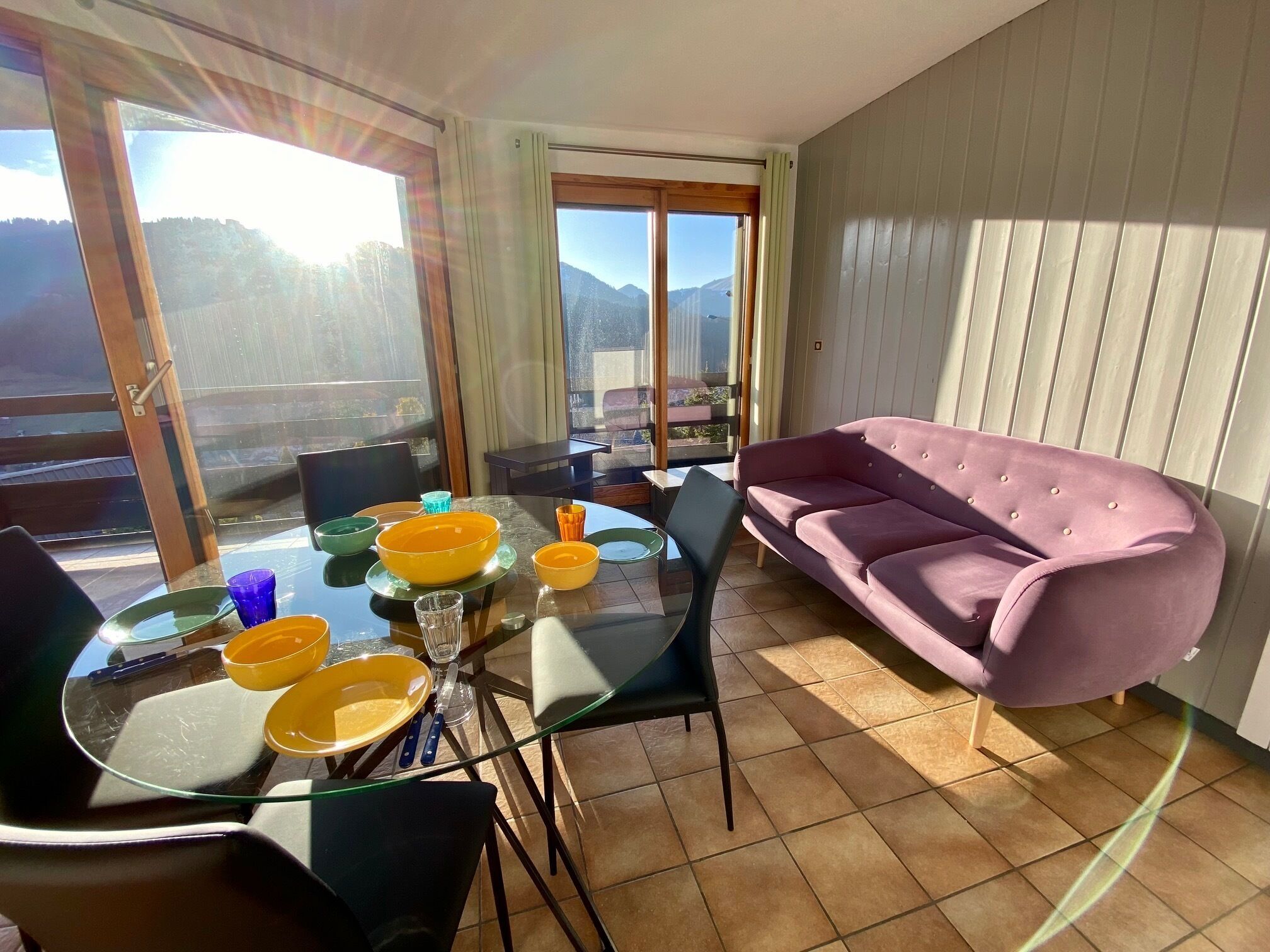 Una sala de estar moderna con una mesa de comedor de vidrio, un sofá rosa y vistas a montañas a través de amplios ventanales.