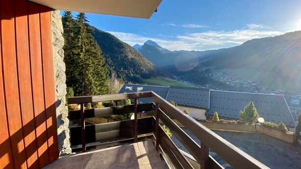 Een balkon met een stenen muur en houten hekwerk met uitzicht op een vallei met bergen en een rivier.