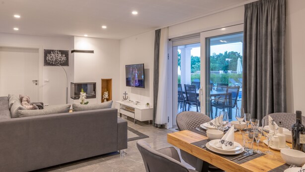 A modern living room with a grey sofa, a wooden dining table set for four, and a TV mounted on the wall.