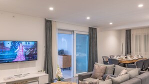 A modern living room with a flat-screen TV, a sofa, a coffee table, and a dining area with chairs and a table set for a meal.