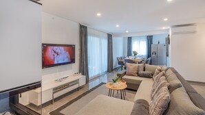 A modern living room with a flat-screen TV, a sofa, a coffee table, and a dining area with a table and chairs.