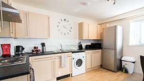 Una cocina moderna con armarios de madera clara, un refrigerador de acero inoxidable, lavadora integrada y un reloj de pared.