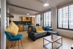 Een moderne woonkamer met een bank, een bijzettafeltje en een barkruk. Achtergrond is een keuken met houten kasten en een fornuis.
