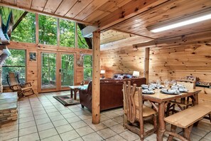 Interior de uma cabana de madeira com uma mesa de jantar posta para quatro pessoas, um sofá de couro e uma lareira.