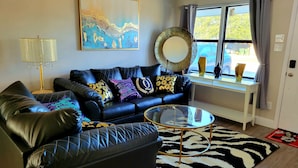Ein Wohnzimmer mit einer schwarzen Ledersofa, einem Glastisch, einem Zebra-Teppich, einem großen abstrakten Gemälde und einem Fenster mit Vorhängen.