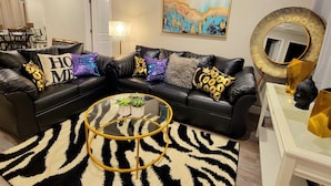 Ein modernes Wohnzimmer mit einem schwarzen Ledersofa, einem Glastisch, einem zebra-gemusterten Teppich und einem goldfarbenen Spiegel.