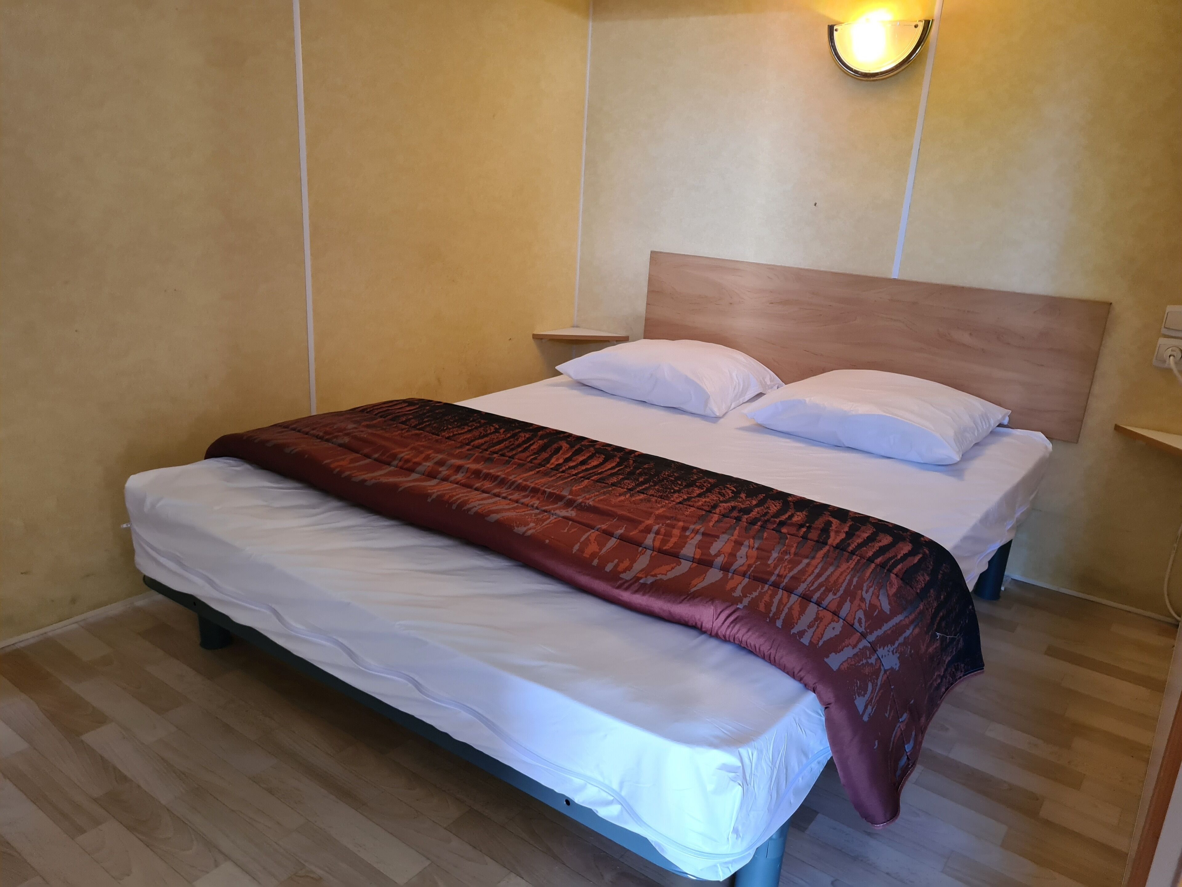 Un lit bien fait, recouvert d’une couverture blanche et marron, avec une tête de lit en bois et une lampe de chevet.