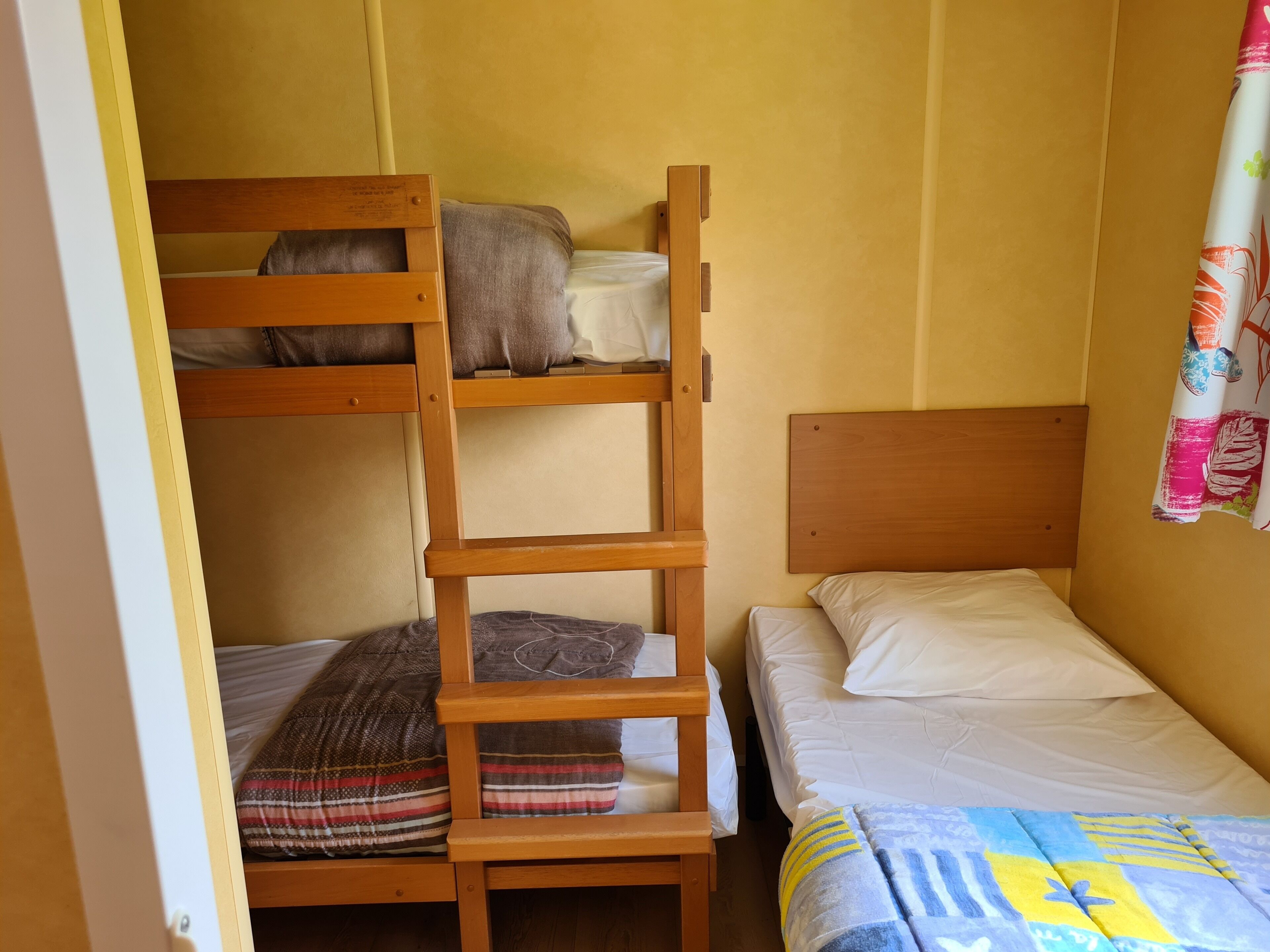 Un lit superposé avec une échelle, un lit simple avec une couverture à motifs et un mur avec un rideau partiellement visible.