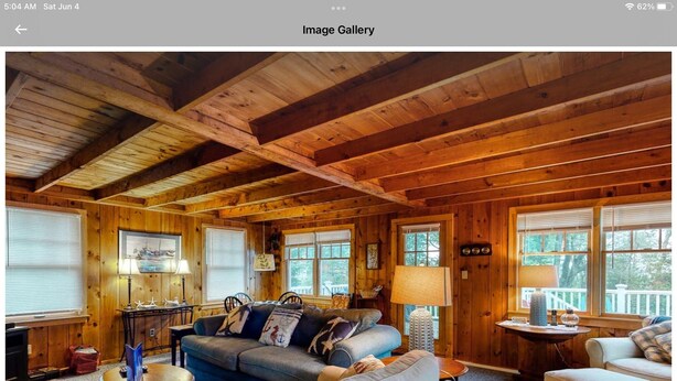 A cozy cabin interior with a wooden ceiling, a sofa, a dining table, and a window with a view of trees. - All Aboard Cottage (East Boothbay)