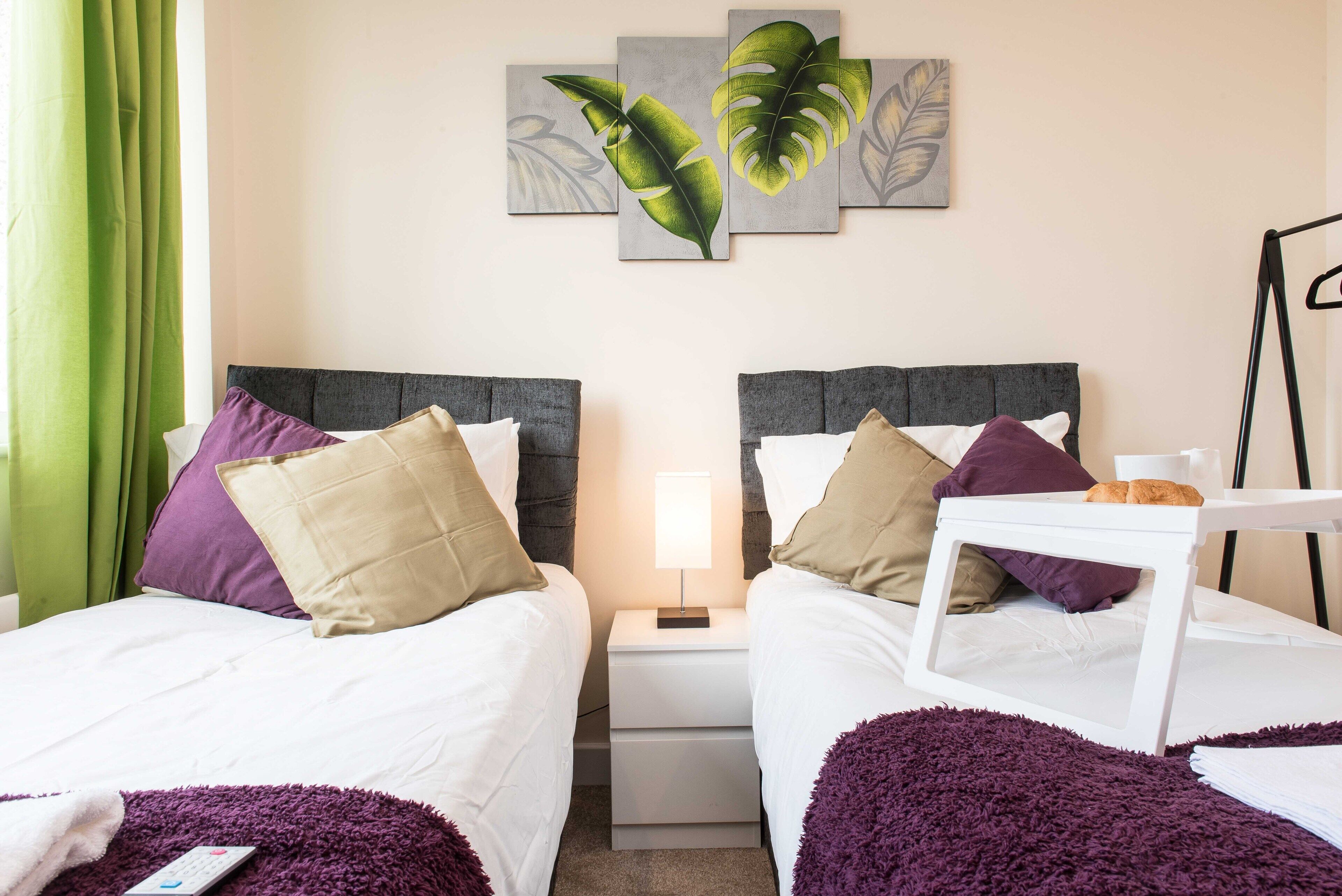 Ein Hotelzimmer mit zwei Betten, einem Nachttisch mit Lampe und einem an der Wand hängenden Kunstwerk mit Blättern.