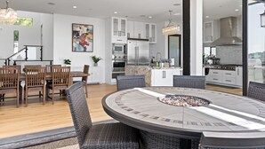 A modern kitchen with white cabinets, a central island with a fire pit, and a dining area with wicker chairs and a wooden table. - 360° HILLTOP VIEW / Ultra Modern / 15min DISNEY (Santa Ana)