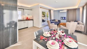 Una sala de estar moderna con comedor, que incluye una cocina con armarios blancos, un conjunto de sofás y un televisor de pantalla plana.