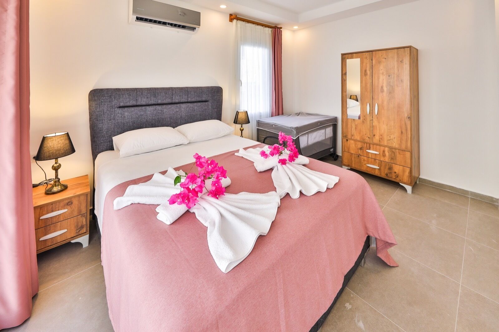Een net opgemaakt bed met roze beddengoed, een houten kledingkast, een nachtkastje met een lamp en een raam met gordijnen.