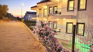 Un edificio residencial de varios pisos con balcones, un camino pavimentado y plantas florecientes a lo largo de la cerca.
