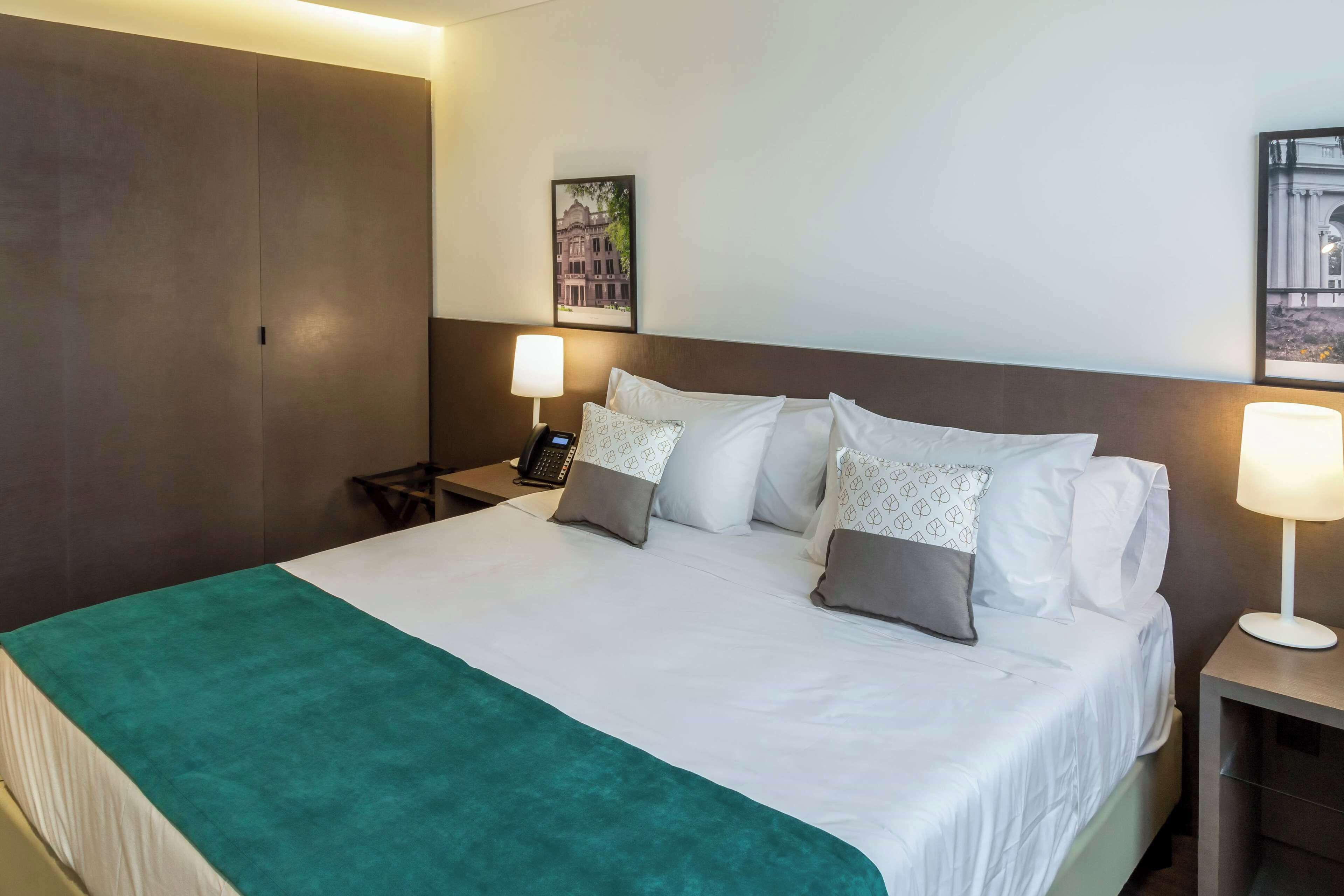 Quarto de hotel com uma cama grande, duas mesas de cabeceira, uma escrivaninha, uma cadeira, um guarda-roupa e duas luminárias de parede.