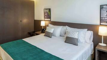 Quarto de hotel com uma cama grande, duas mesas de cabeceira, uma escrivaninha, uma cadeira, um guarda-roupa e duas luminárias de parede.