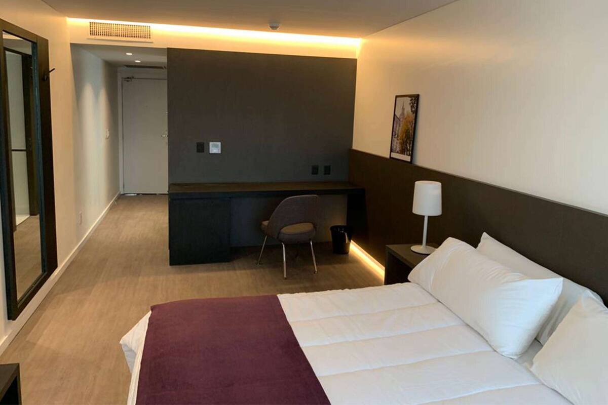 A hotel room with a bed, a desk, a chair, and a nightstand with a lamp.