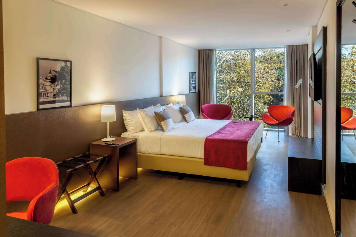 A modern hotel room with a large bed, a desk, two red chairs, a TV, and a window with a view of trees.