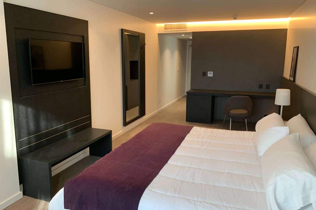 A modern hotel room with a large bed, a flat-screen TV mounted on the wall, a desk with a chair, and a small seating area.