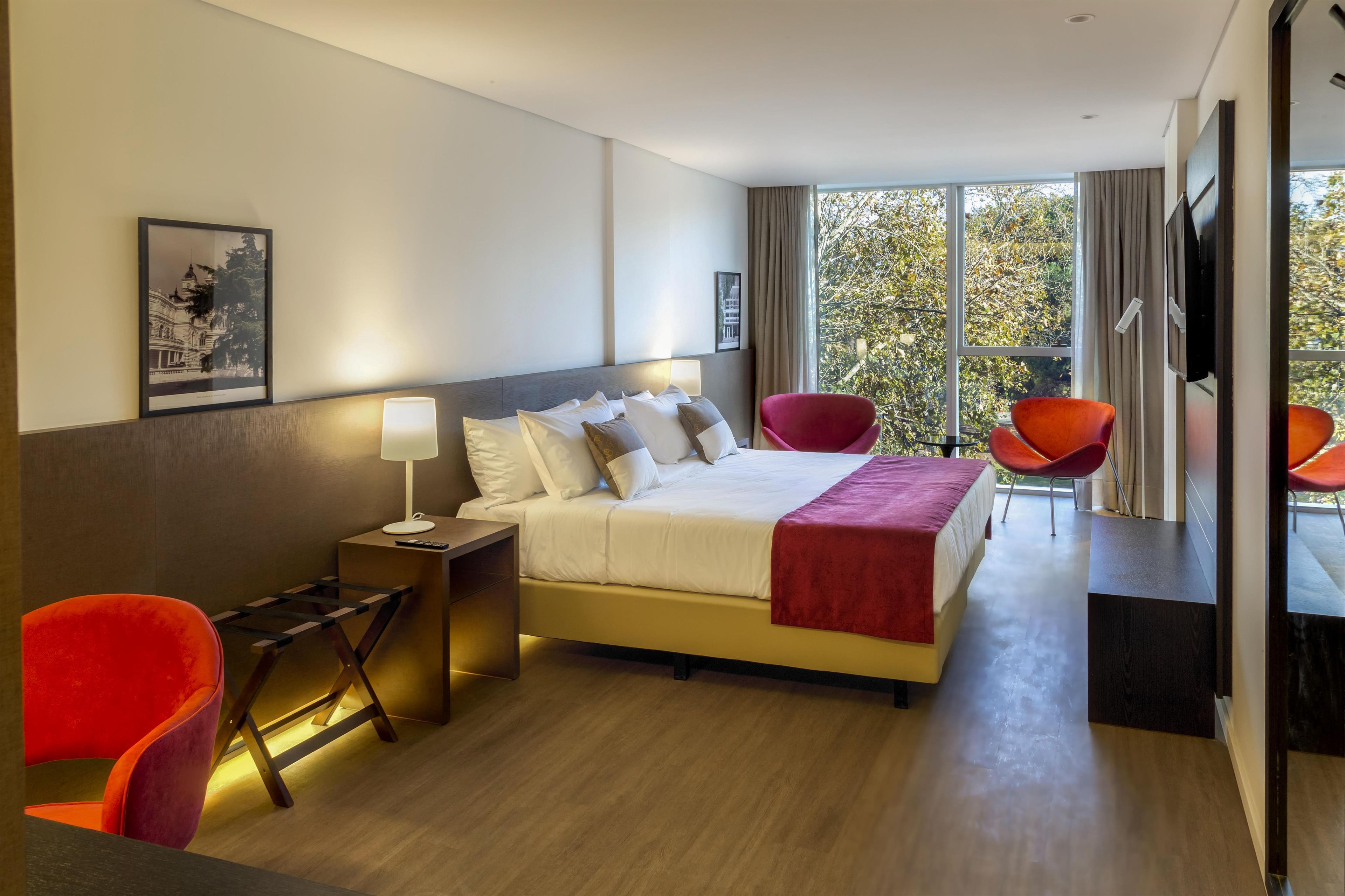 Habitación de hotel con cama, mesita de noche, lámpara, dos sillones y un amplio ventanal con vistas a árboles.