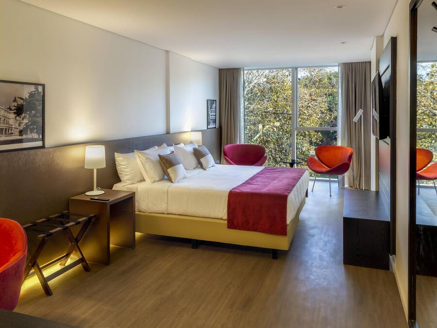 A hotel room with a bed, bedside table, lamp, two chairs, and a large window with a view of trees.