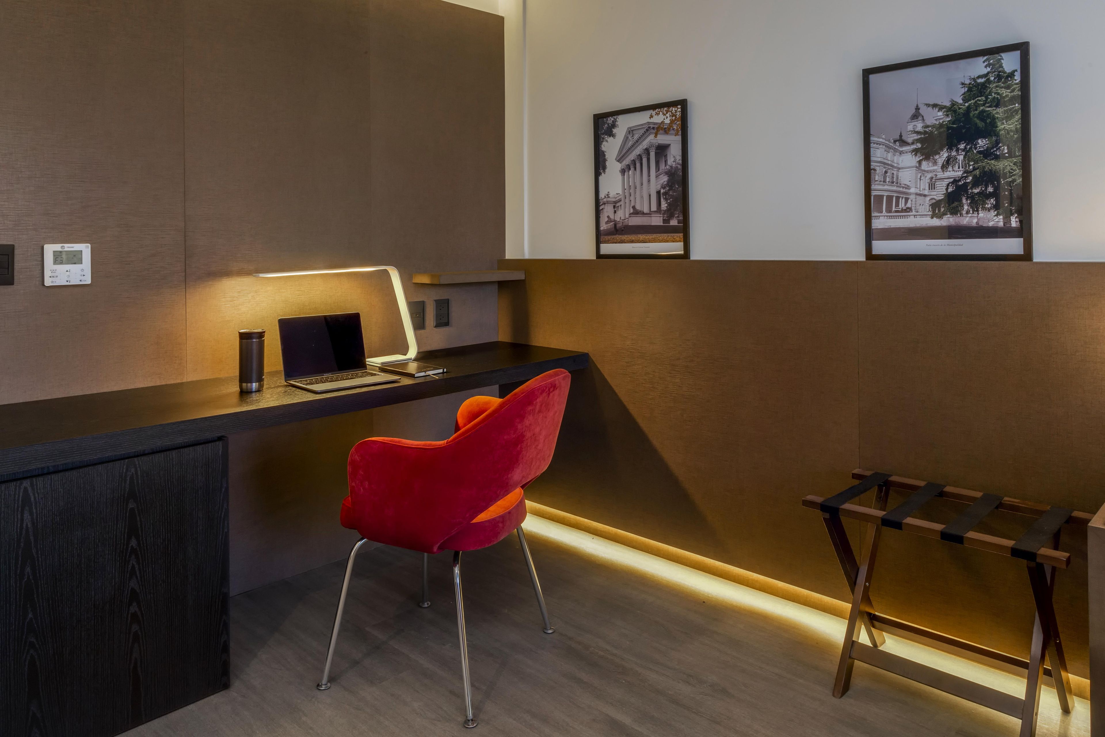 A modern workspace with a desk, laptop, and a red chair. There are framed pictures on the wall and a small table with a lamp.