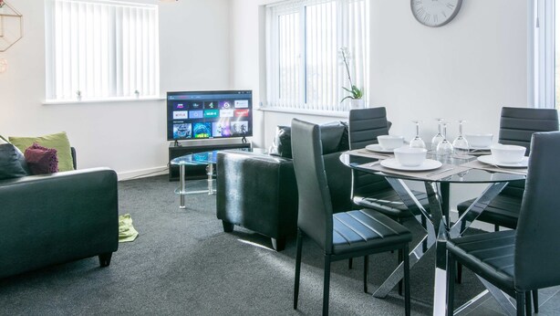 A modern living room with a glass dining table, black chairs, a flat-screen TV, and a sofa with cushions. - Levon House, Coventry - 2 Bedroom Apartment (Coventry)