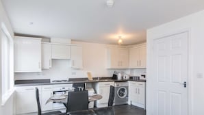 A compact kitchen with white cabinets, a dining table with chairs, a washing machine, and a small seating area with a sofa and a chair. - Levon House, Coventry - 2 Bedroom Apartment (Coventry)