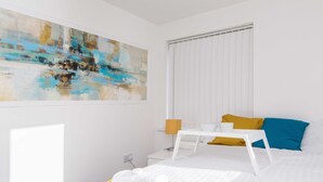A modern hotel room with a large abstract painting, a bed with white and yellow bedding, a bedside table with a lamp, and a window with blinds. - Levon House, Coventry - 2 Bedroom Apartment (Coventry)