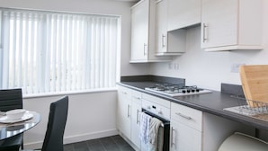 A compact kitchen with white cabinets, a gas stove, and a dining area with a table and chairs. - Levon House, Coventry - 2 Bedroom Apartment (Coventry)