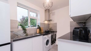 A compact kitchen with a built-in washer and dryer, a window with a view of greenery, and a modern stove. - Michaelmas, Coventry - 2 Bed House Apartment (Coventry)