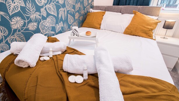 A neatly made bed with white and mustard bedding, a tray with a bun and a mug, and a white tray with a bottle and two round objects. - Michaelmas, Coventry - 2 Bed House Apartment (Coventry)