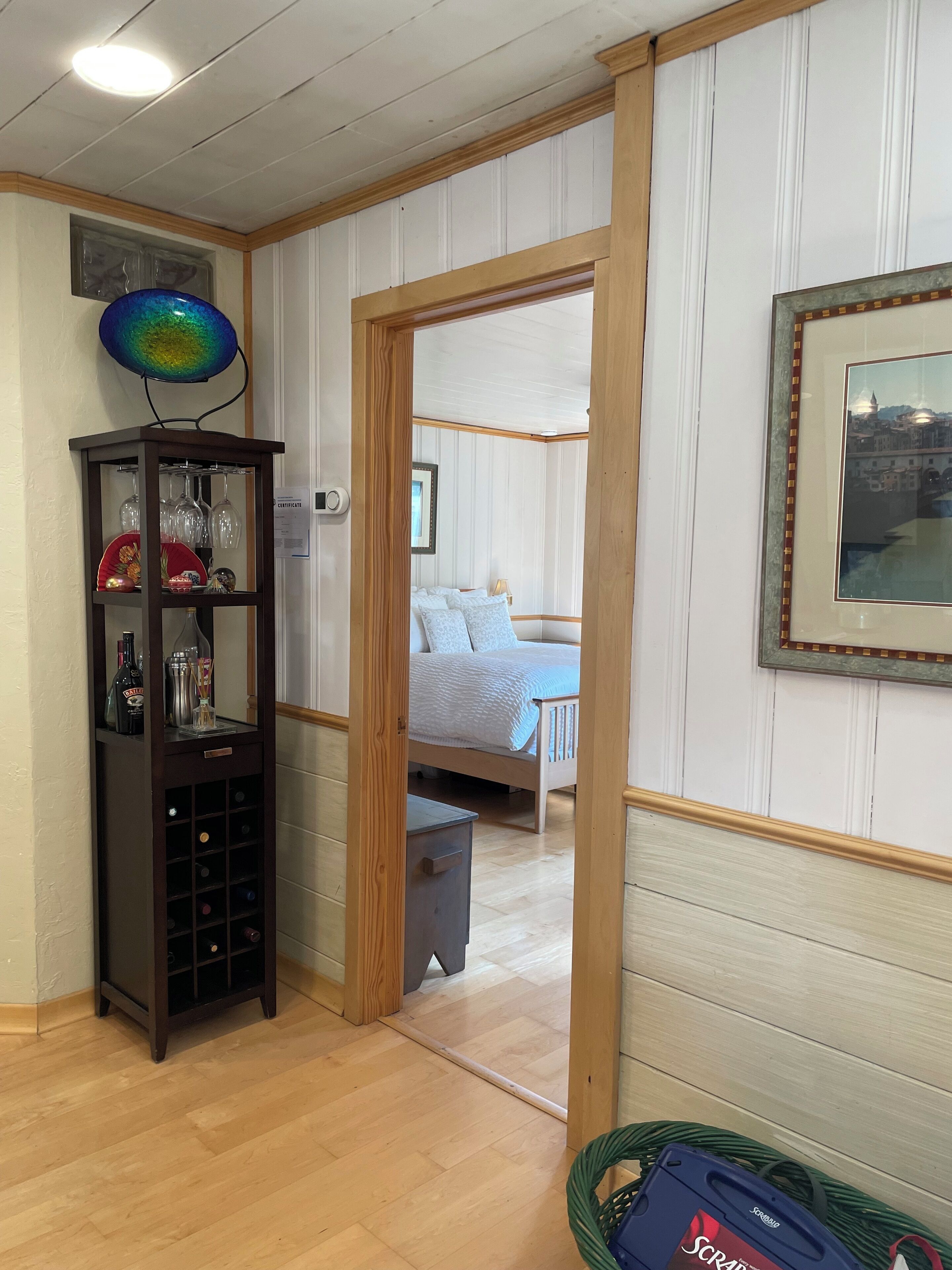 Une pièce avec un plancher en bois, une armoire sombre également en bois contenant des bouteilles de vin, un tableau encadré accroché au mur et un lit recouvert d’une couverture bleue.