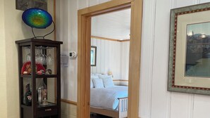 Una habitación con piso de madera, un mueble de madera oscura con botellas de vino, un cuadro en la pared y una cama con una manta azul.
