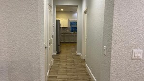 A narrow hallway with wooden flooring leading to a kitchen area with white cabinets and a refrigerator.