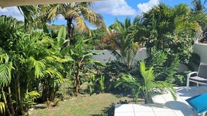 Un patio con suelo de baldosas blancas, una silla de descanso y una mesita. Se observan diversas plantas tropicales y una palmera.