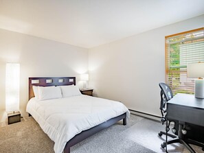 Habitación de hotel con cama, mesita de noche, lámpara, escritorio, silla y ventana con persianas.