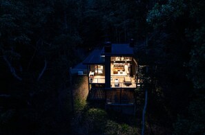 A two-story wooden cabin with a deck, surrounded by trees.